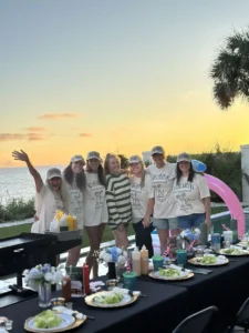 Group of women at beachside hibachi dinner at sunset, wearing matching hats and shirts