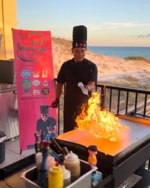 Hibachi chef performing fire trick on grill by the beach with MyHibachi banner in the background
