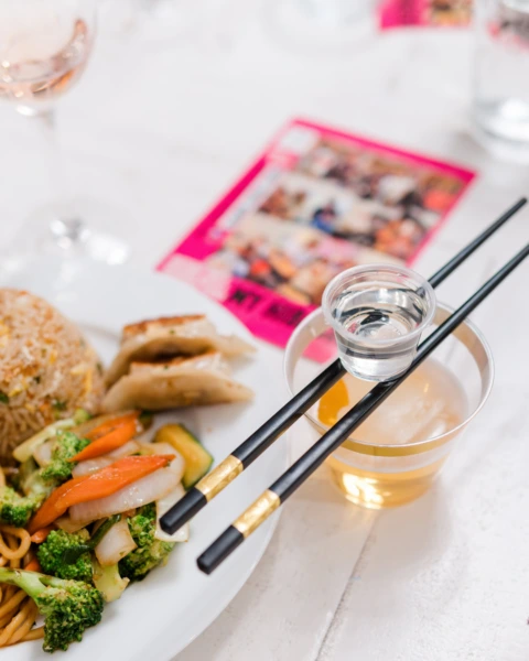Hibachi plate with noodles, dumplings, veggies and drinks with chopsticks balancing a sake shot