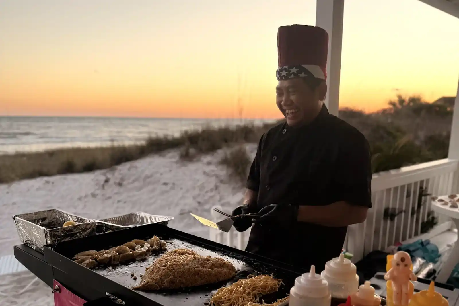 Hibachi chef cooking by the beach at sunset with noodles and grilled meats on grill